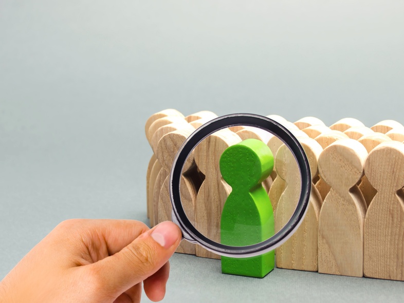 En hand håller ett förstoringsglas över en grön träfigur bland flera beige träfigurer.
