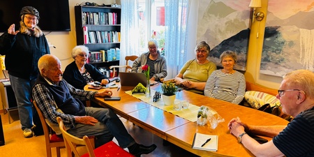 En grupp äldre personer sitter runt ett bord i ett ljust rum, en person står och pratar.