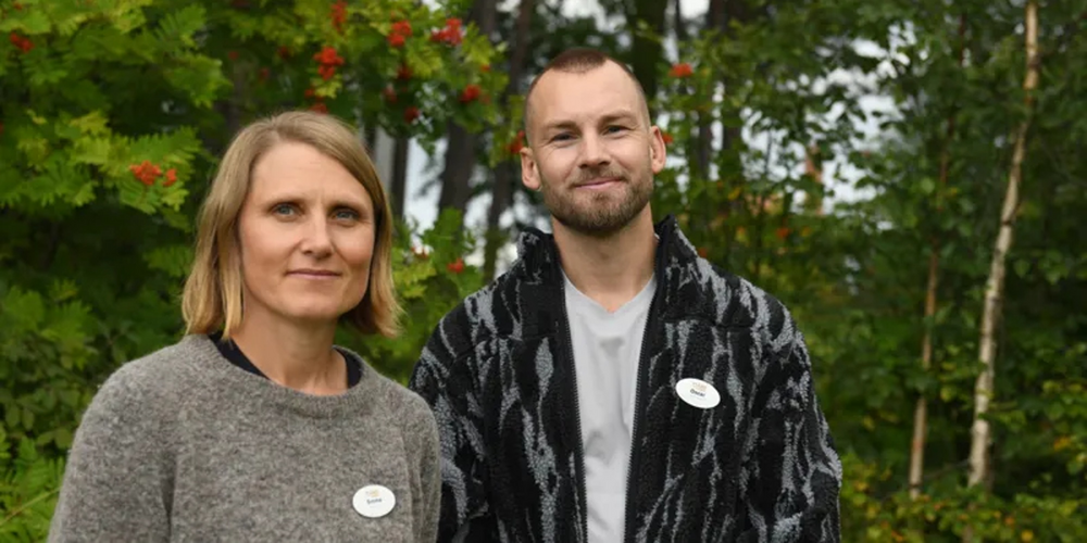 Två personer står utomhus framför grönskande träd och bär namnskyltar.
