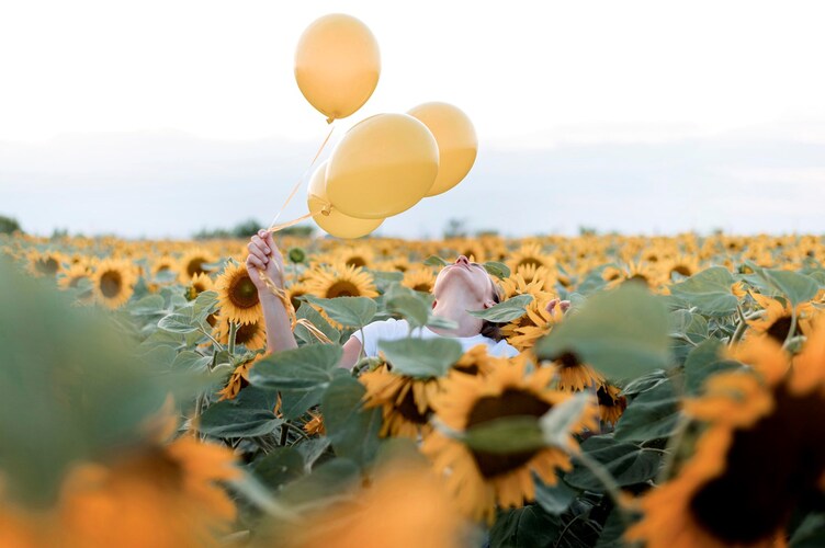 En person står i ett fält av solrosor och håller upp tre gula ballonger mot himlen.