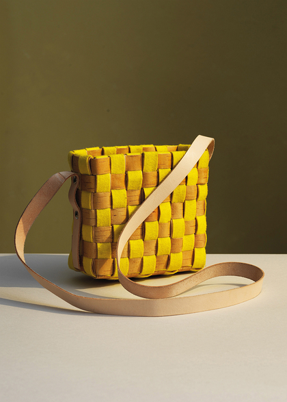 En liten väska med flätad design i gult och brunt, med en lång, ljus axelrem, står på ett bord mot en olivgrön bakgrund.