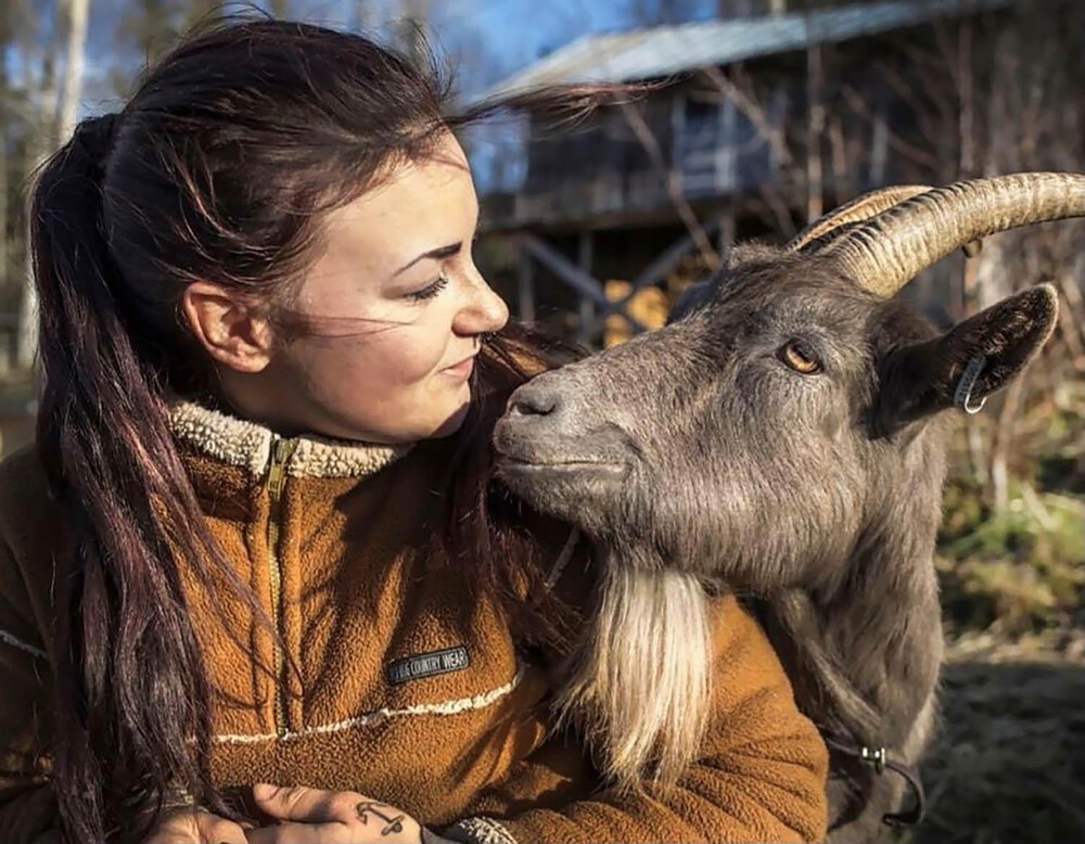 En kvinna i brun fleecejacka tittar leende på en get som står nära henne utomhus.