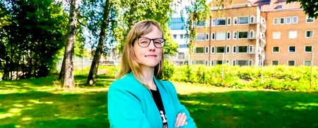 Person står utomhus och tittar in i kameran. Hen har glasögon och längre hår med snedlugg. Solen skiner och det är grönskande bakgrund.