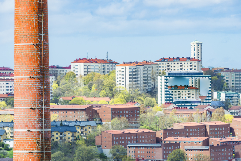Stadsbild med röda tegelhus, höghus och en stor skorsten i förgrunden under en blå himmel.