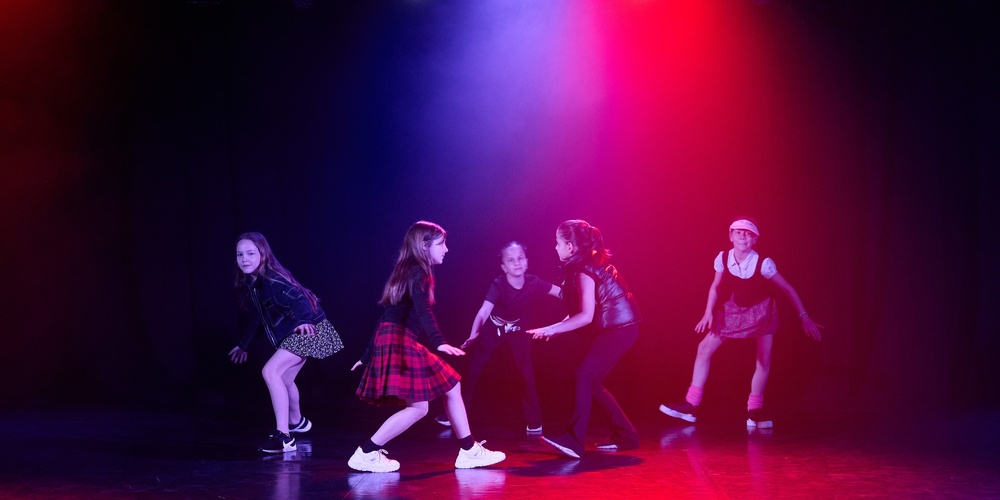 Fem unga personer dansar på en scen med färgglada ljus i bakgrunden.