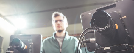 En person står suddigt i bakgrunden bakom två filmkameror i en studio med stark belysning.