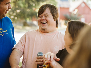 Tre personer står utomhus och skrattar tillsammans, en håller i en mikrofon och en annan i en gitarr.