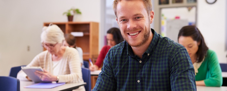 Leende man sitter vid skolbänk i klassrum, skriver i anteckningsbok. Andra vuxna studenter i bakgrunden.