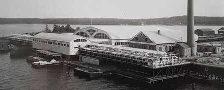 Svartvit bild av en industribyggnad vid vattnet med stor skorsten och bryggor.