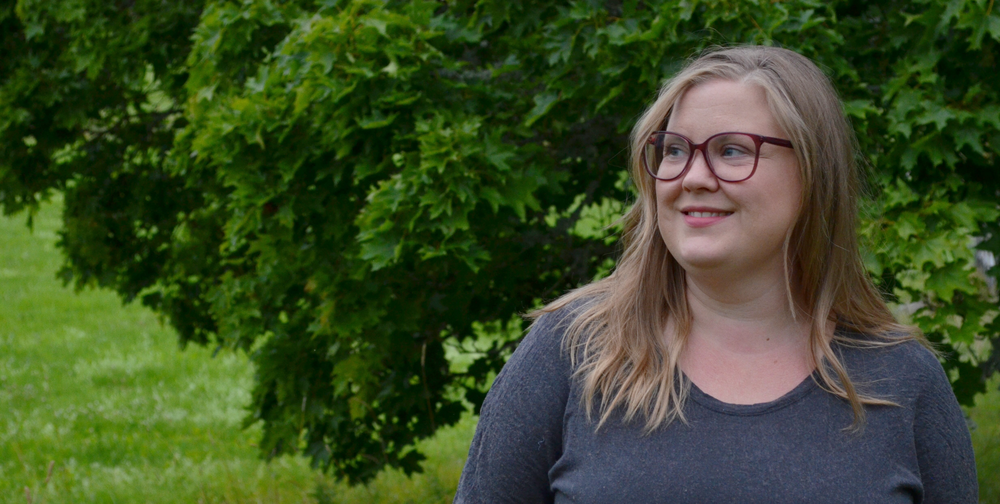 Person med långt blont hår står utomhus framför gröna träd, ansiktet är suddat.