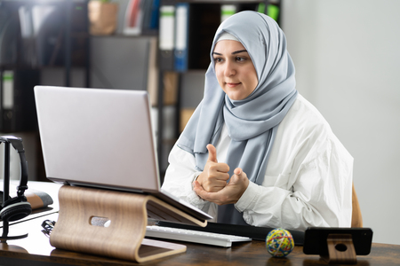 Kvinna med hijab sitter vid ett skrivbord och deltar i ett videosamtal på en bärbar dator.