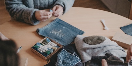 Tre personer sitter runt ett bord och arbetar med tygbitar och broderimaterial.