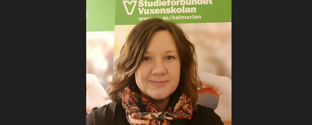 Kvinna med brunt hår och mönstrad halsduk framför en grön skylt med texten 'Studieförbundet Vuxenskolan'.