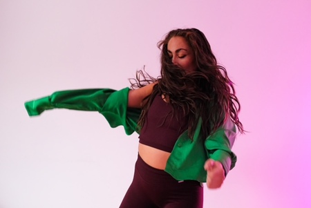 Kvinna dansar energiskt i grönt plagg mot ljus bakgrund med rosa ton.