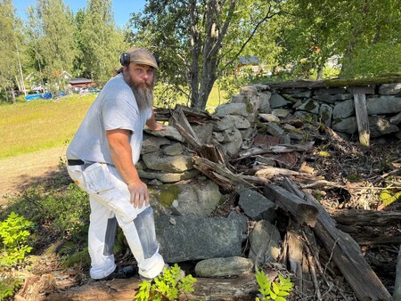 En person undersöker en gammal, delvis raserad stenmur i en solig skogsmiljö.