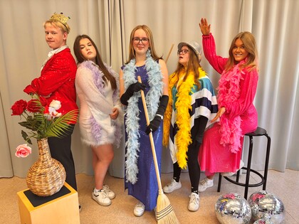 Fem personer i färgglada utklädnader poserar framför ett draperi med rekvisita som fjäderboor, diskokulor och en kvast.