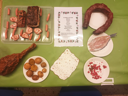 Ett bord med olika maträtter, en meny i mitten och tallrikar med kött, bröd och bär på en grön duk.