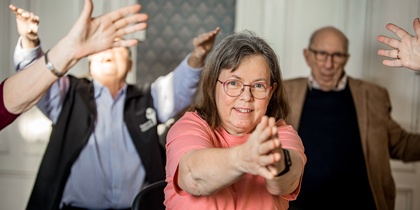 En grupp äldre personer gör gymnastik tillsammans inomhus, en kvinna i rosa tröja i fokus.