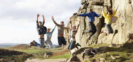 Sex personer hoppar glada i luften vid en klippa i ett naturskönt landskap.