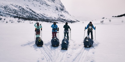 Fyra personer med pulkor står på skidor i ett snötäckt fjällandskap med berg i bakgrunden.