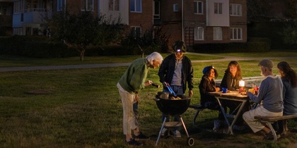 En grupp människor grillar och sitter vid ett bord upplyst av batterilampa utomhus på kvällen framför ett flerbostadshus.