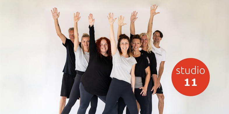 En grupp glada vuxna i träningskläder poserar med armarna uppsträckta framför en vit bakgrund.