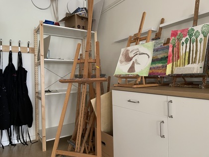 Ett konstnärsrum med stafflier, målningar, svarta förkläden och hyllor.