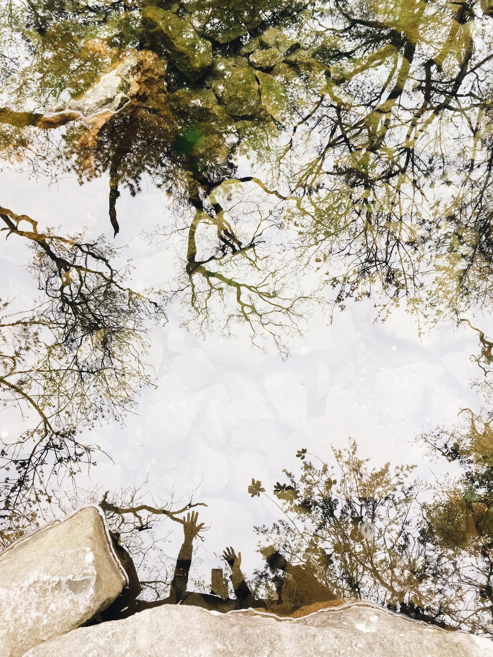 Reflektion av träd och två personer med uppsträckta armar i en vattenyta, stenar i förgrunden.