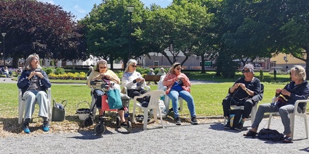 Sex personer sitter utomhus i en park och stickar tillsammans i solskenet.