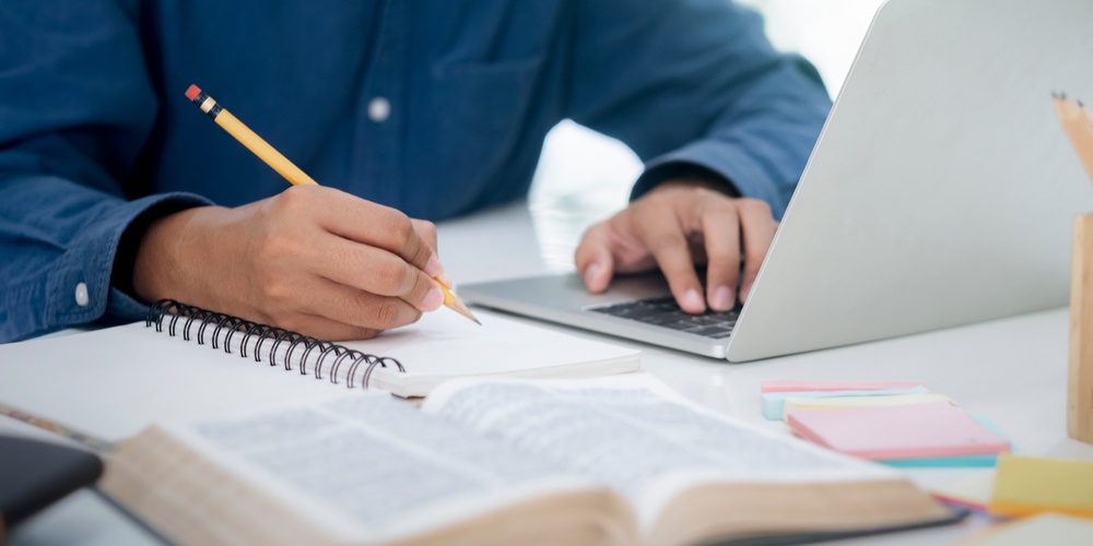 En person skriver i en anteckningsbok vid ett skrivbord med en laptop och en uppslagen bok.