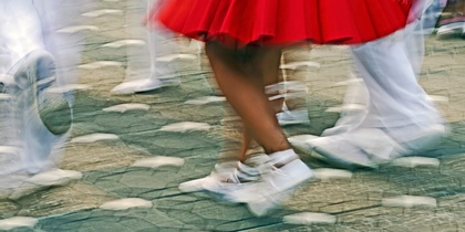 Personer dansar i rörelse, klädda i vita kläder och röd kjol, på en stenlagd gata.