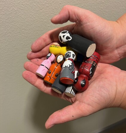 Två händer håller flera små, handmålade träfigurer som föreställer olika karaktärer.