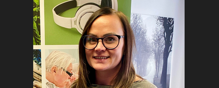 Kvinna med glasögon och brunt hår står inomhus framför färgglada affischer.