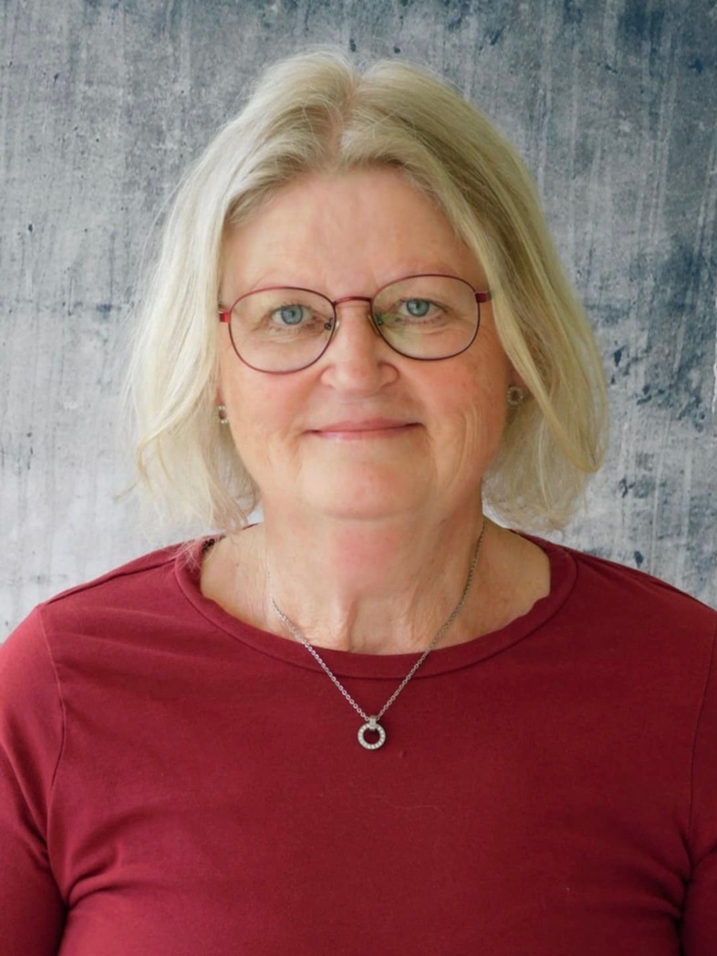 Lena Larsson med blont hår, röd tröja och halsband står mot en grå bakgrund.