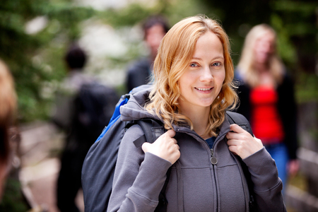 Ung kvinna med ryggsäck och grå huvtröja ler utomhus, suddiga personer i bakgrunden.