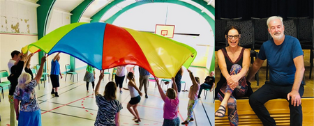 Grupp människor leker med en färgglad fallskärm i en gympasal, två vuxna sitter på en scen bredvid.