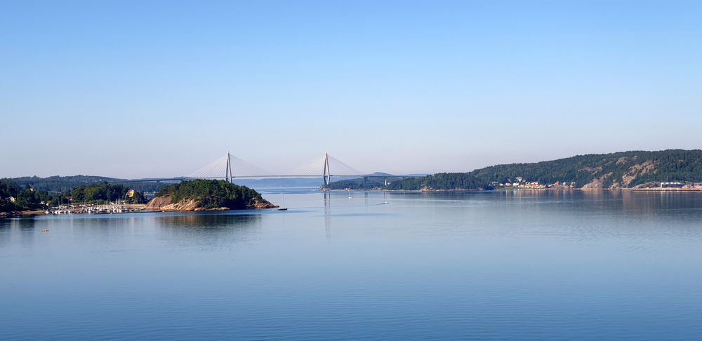 Utsikt över en lugn fjord med en bro i bakgrunden och skogsklädda kullar på sidorna.