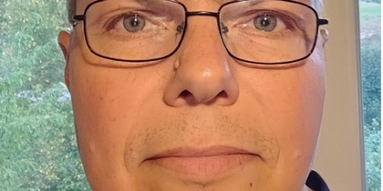 Man med glasögon tittar rakt in i kameran, inomhus med fönster och grönska i bakgrunden.
