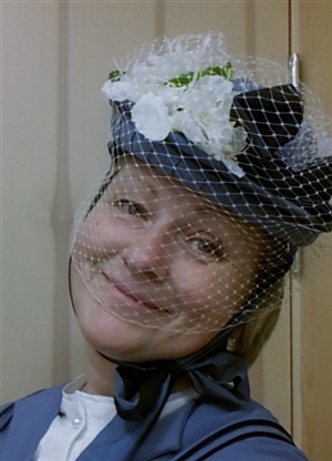 Person med ljusblå kavaj och vit blus bär en hatt med vita blommor och nätdekoration.