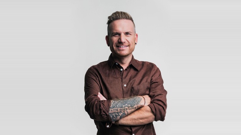 Man med tatuerad arm står med armarna i kors och ler mot kameran, mot en ljus bakgrund.