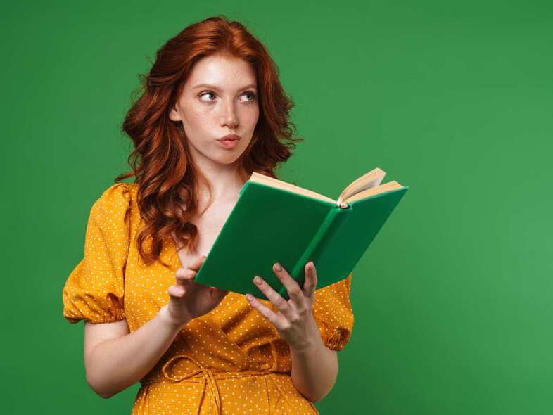 En kvinna i gul tunika står framför en grön vägg och läser en grön bok.