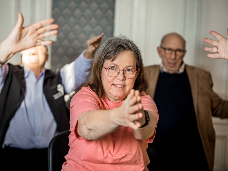 En grupp äldre personer gör gymnastik tillsammans inomhus, en kvinna i rosa tröja i fokus.