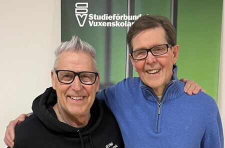 Två personer står tillsammans framför en skylt med texten 'Studieförbundet Vuxenskolan'.