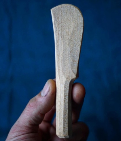 En hand håller en ofärdig smörkniv av trä mot en blå bakgrund.