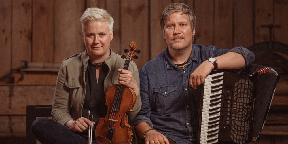 Två personer sitter bredvid varandra, en håller en fiol och den andra ett dragspel, i en träpanelklädd miljö.