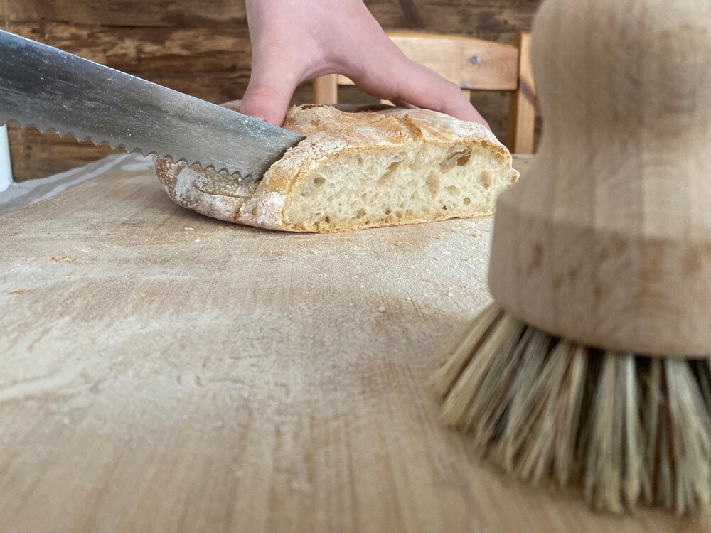 En hand skär en brödbit med en brödkniv på en träskärbräda, en borste syns i förgrunden.