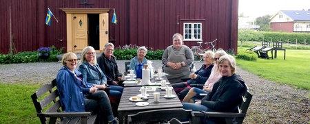 En grupp vuxna sitter runt ett bord utomhus framför ett rött trähus och fikar tillsammans.