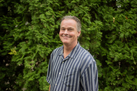 Man med randig skjorta står utomhus framför en grön häck och ler mot kameran.