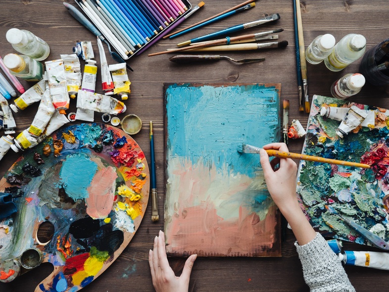 En person målar abstrakt konst på en duk med pensel, omgiven av färgtuber, penslar och färgpaletter på ett bord.