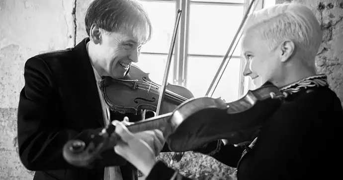 Två personer spelar violin tillsammans och ler mot varandra i ett ljust rum.